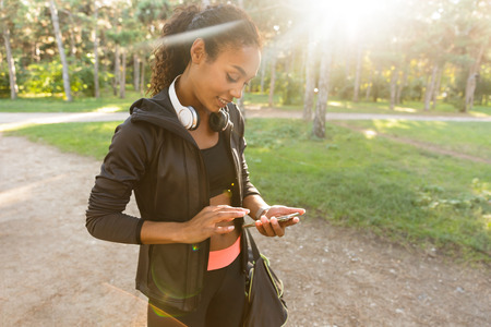 Image of pleased woman 20s wearing black tracksuit and headphones using smartphone in green park