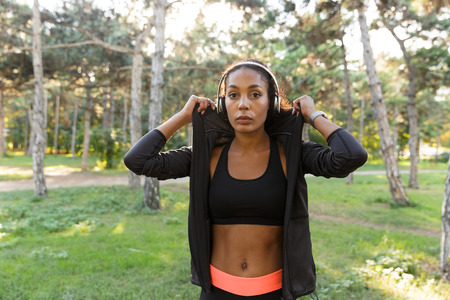 Portrait of sportswoman 20s wearing black tracksuit and headphones walking through green park