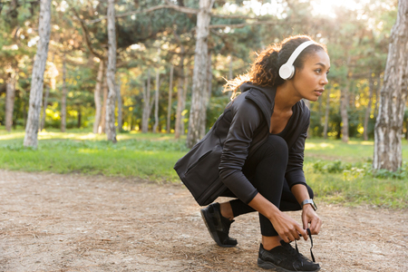 Image of brunette sportswoman 20s wearing black tracksuit tying her shoelaces while running through green parkの写真素材