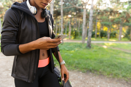 Cropped Image of athletic woman 20s wearing black tracksuit and headphones using smartphone in green parkの写真素材