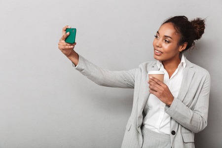 Attractive young african business woman standing isolated over gray background, holding cup of takeaway coffee, taking a selfieの写真素材