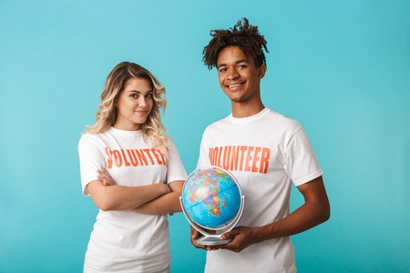 Happy confident multiethnic couple wearing volunteers t-shirt standing isolated over blue background, holding a globeの写真素材