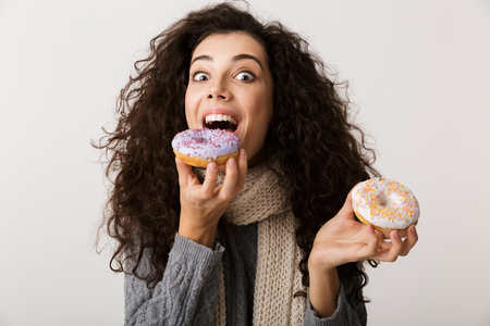 Attractive young woman wearing winter scarf showing sweet donuts while standing isolated over white backgroundの写真素材
