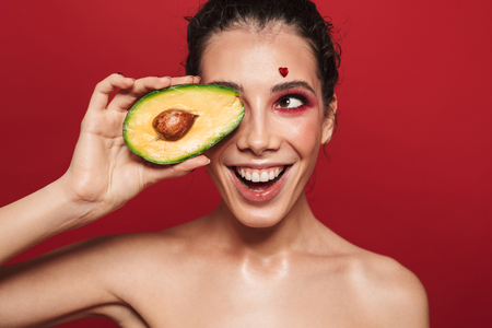 Beauty portrait of an attractive young woman wearing makeup standing isolated over red background, posing, holding sliced avocadoの写真素材