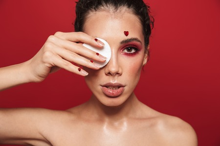 Beauty portrait of an attractive young topless woman wearing makeup standing isolated over red background, posing, holding cotton padsの写真素材