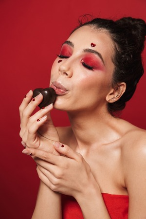 Beauty portrait of an attractive young topless woman wearing makeup standing isolated over red background, holding heart shaped chocolate candyの写真素材
