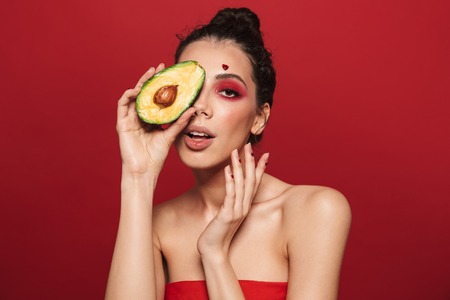 Beauty portrait of an attractive young woman wearing makeup standing isolated over red background, posing, holding sliced avocadoの写真素材