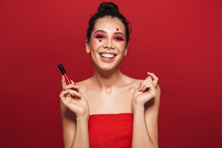 Beauty portrait of an attractive young topless woman wearing makeup standing isolated over red background, posing, holding lip glossの写真素材
