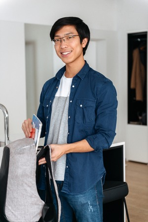 Smiling asian man packing his backpack, ready to leave apartmentの写真素材
