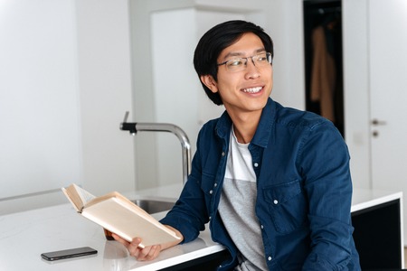 Smiling asian man reading book while sitting on a kitchen at homeの写真素材