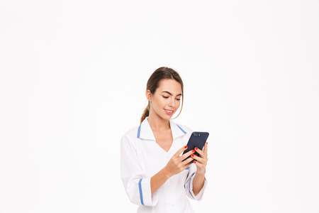 Confident young woman doctor wearing uniform standing isolated over white background, using mobile phoneの写真素材