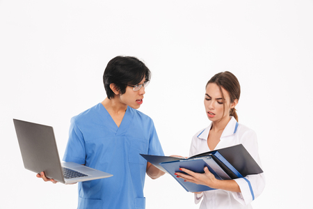 Serious doctors couple wearing uniform standing isolated over white background, working with laptop computer and x-ray photographの写真素材