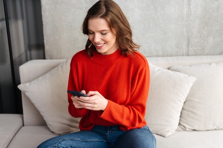 Cheerful young casually dressed woman sitting on a couch at home, holding mobile phoneの写真素材