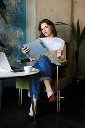 Picture of pretty beautiful young woman sitting in cafe indoors work with laptop holding documents.の写真素材