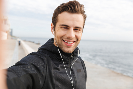 Image of smiling sportsman 30s in black sportswear and earphones taking selfie photo on mobile phone while walking at seasideの写真素材