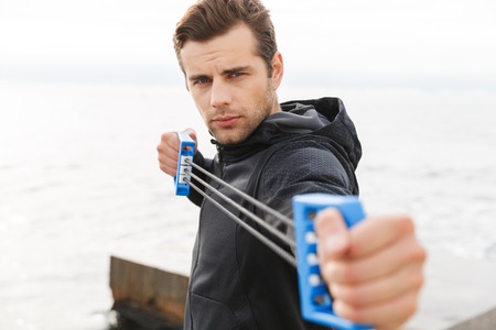 Handsome confident young sportsman working out at the beach, using chest expander