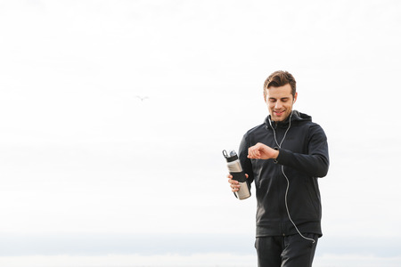 Image of satisfied sportsman 30s in black sportswear and earphones working out and running by...