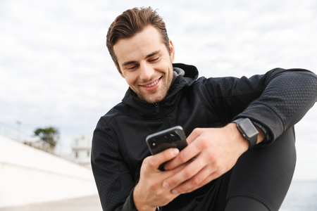 Image of sportive man 30s in black sportswear using smartphone while sitting on boardwalk at seasideの写真素材