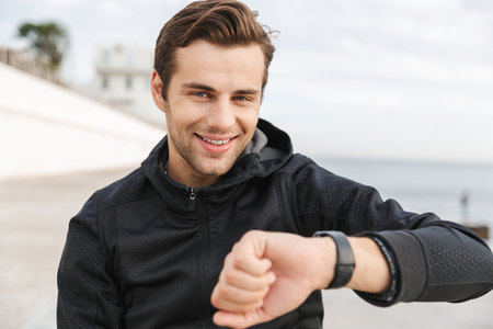 Image of joyful guy 30s in black sportswear looking at wrist watch while sitting on boardwalk at seasideの写真素材