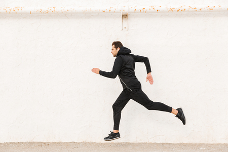 Image of caucasian man 30s in black sportswear and earphones running along white wall outdoorの写真素材