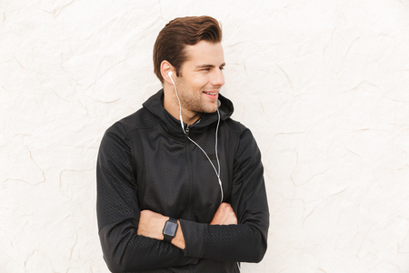 Image of attractive sportsman 30s in black sportswear and earphones standing over white wall outdoorの写真素材