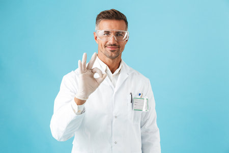 Confident man doctor wearing uniform standing isolated over blue background giving the OK signの写真素材
