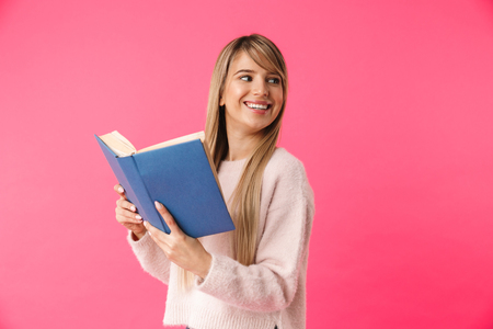 Cheerful young blonde girl standing isolated over pink background, reading a bookの写真素材