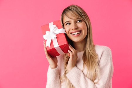 Cheerful young blonde girl standing isolated over pink background, showing gift boxの写真素材