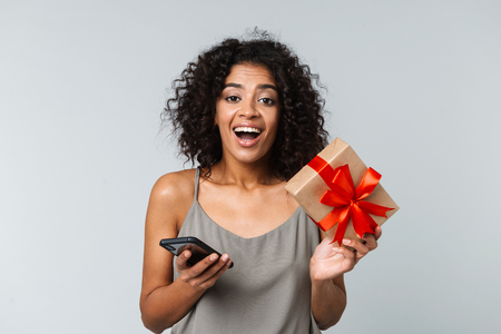 Happy young african woman casually dressed standing isolated over gray background, using mobile phone, showing gift boxの写真素材