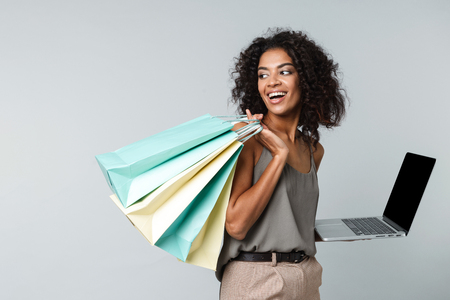 Happy young african woman casually dressed standing isolated over gray background, holding laptop computer, carrying shopping bagsの写真素材