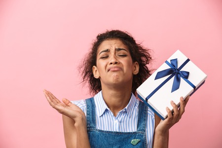 Beautiful disappointed young african woman holding gift box over pink backgroundの写真素材