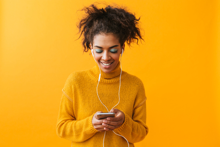 Cheerful african woman wearing sweater standing isolated over yellow background, listening to music with earphones, holding mobile phoneの写真素材