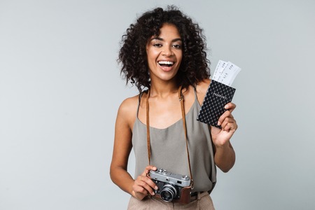 Happy young african woman casually dressed standing isolated over gray background, holding photo camera, showing passport with flight ticketsの写真素材