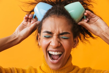 Cheerful african woman wearing sweater holding cups on her head isolated over yellow background