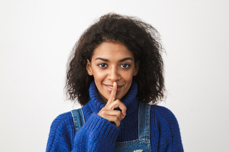 Close up of a pretty young african woman wearing sweater standing isolated over white background, showing silence gestureの写真素材