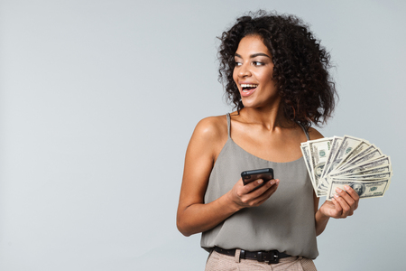 Happy young african woman standing isolated over gray background, holding bunch of money banknotes, using mobile phoneの写真素材