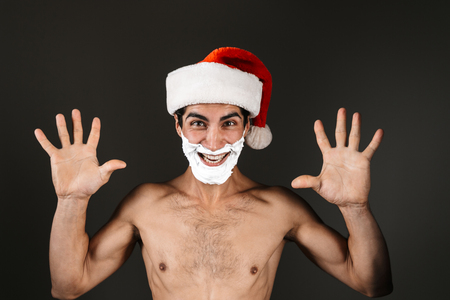 Close up of an excited shirtless man wearing santa claus hat standing isolated over black background, with raised handsの写真素材