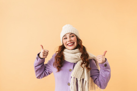 Photo of positive woman 20s in winter clothes showing thumbs up while standing isolated over beige backgroundの写真素材