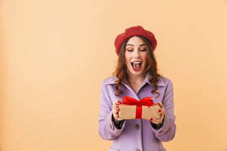 Image of gorgeous woman 20s with long hair smiling and holding present box standing isolated over beige backgroundの写真素材