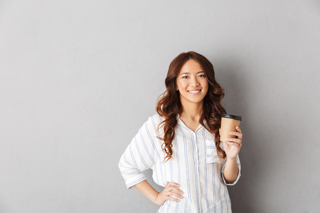 Cheerful asian woman standing isolated over gray background, drinking coffeeの写真素材