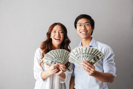Happy asian business couple standing isolated over gray background, holding money banknotesの写真素材