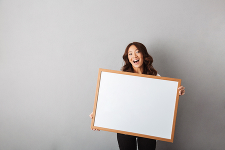 Cheerful asian woman standing isolated over gray background, showing blank boardの写真素材