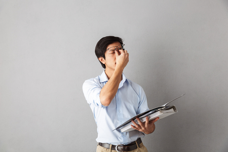 Tired asian man standing isolated over gray background, carrying folders with documentsの写真素材