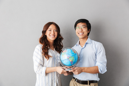 Cheerful asian couple standing isolated over gray background, holding earth globeの写真素材