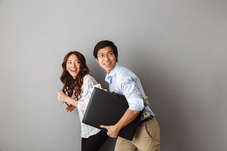 Happy asian couple running away isolated over gray background, holding briefcase full of money banknotesの写真素材