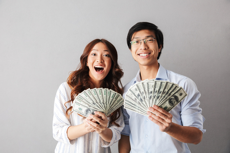 Happy asian business couple standing isolated over gray background, holding money banknotesの写真素材