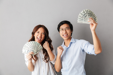 Cheerful asian couple standing isolated over gray background, holding money banknotesの写真素材