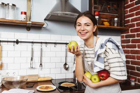 Happy young woman standing at the kitchen at home, holding applesの写真素材
