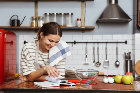 Happy young woman standing at the kitchen at home, reading recipe bookの写真素材