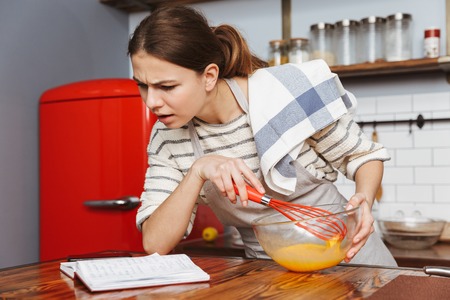 Serious young woman standing at the kitchen at home, mixing eggs in a bowlの写真素材